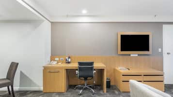 A hotel room with a wooden desk, a black office chair, a television mounted on the wall, and a patterned carpet.