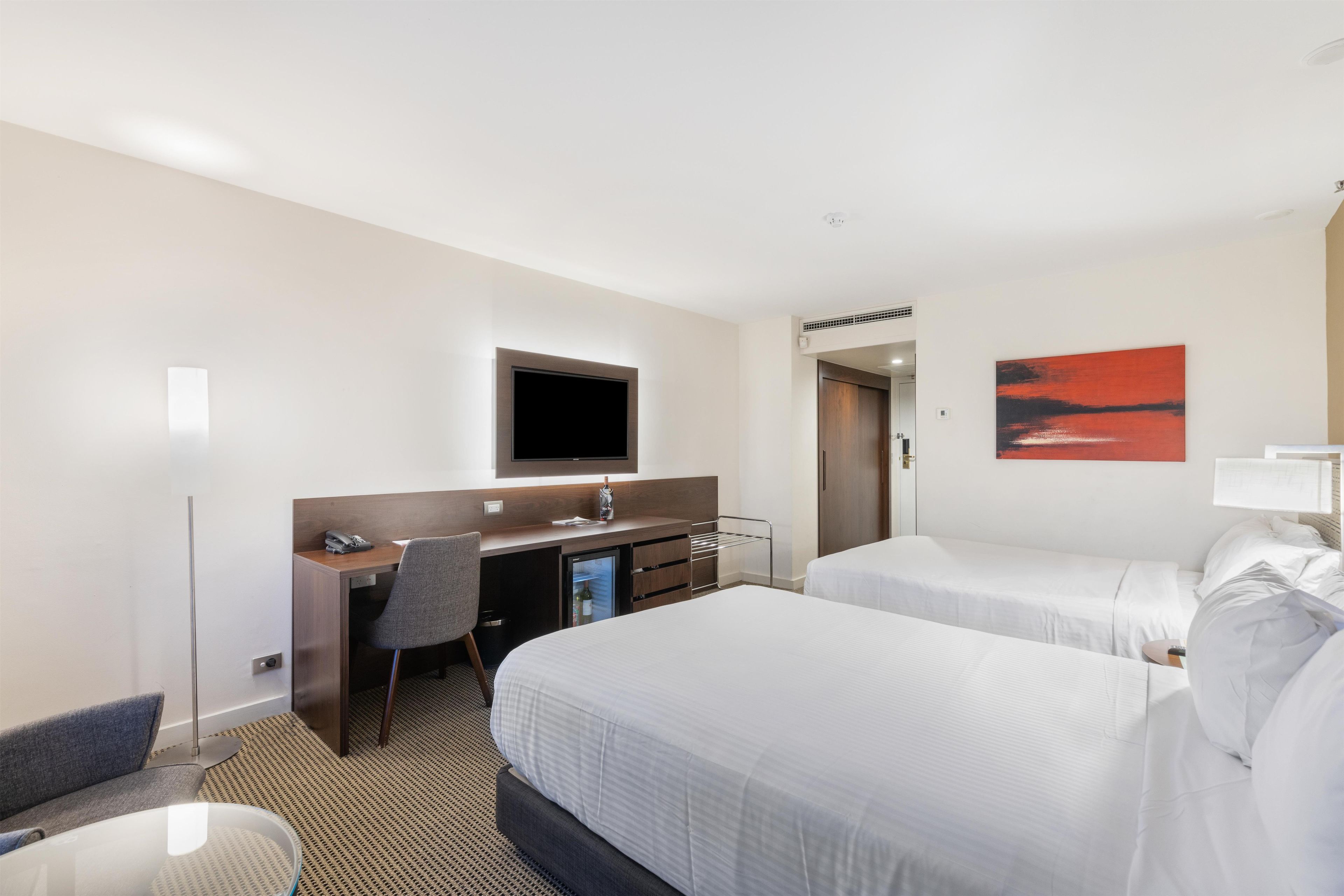 Een hotelkamer met twee bedden, een bureau met stoel, een televisie en een schilderij aan de muur.