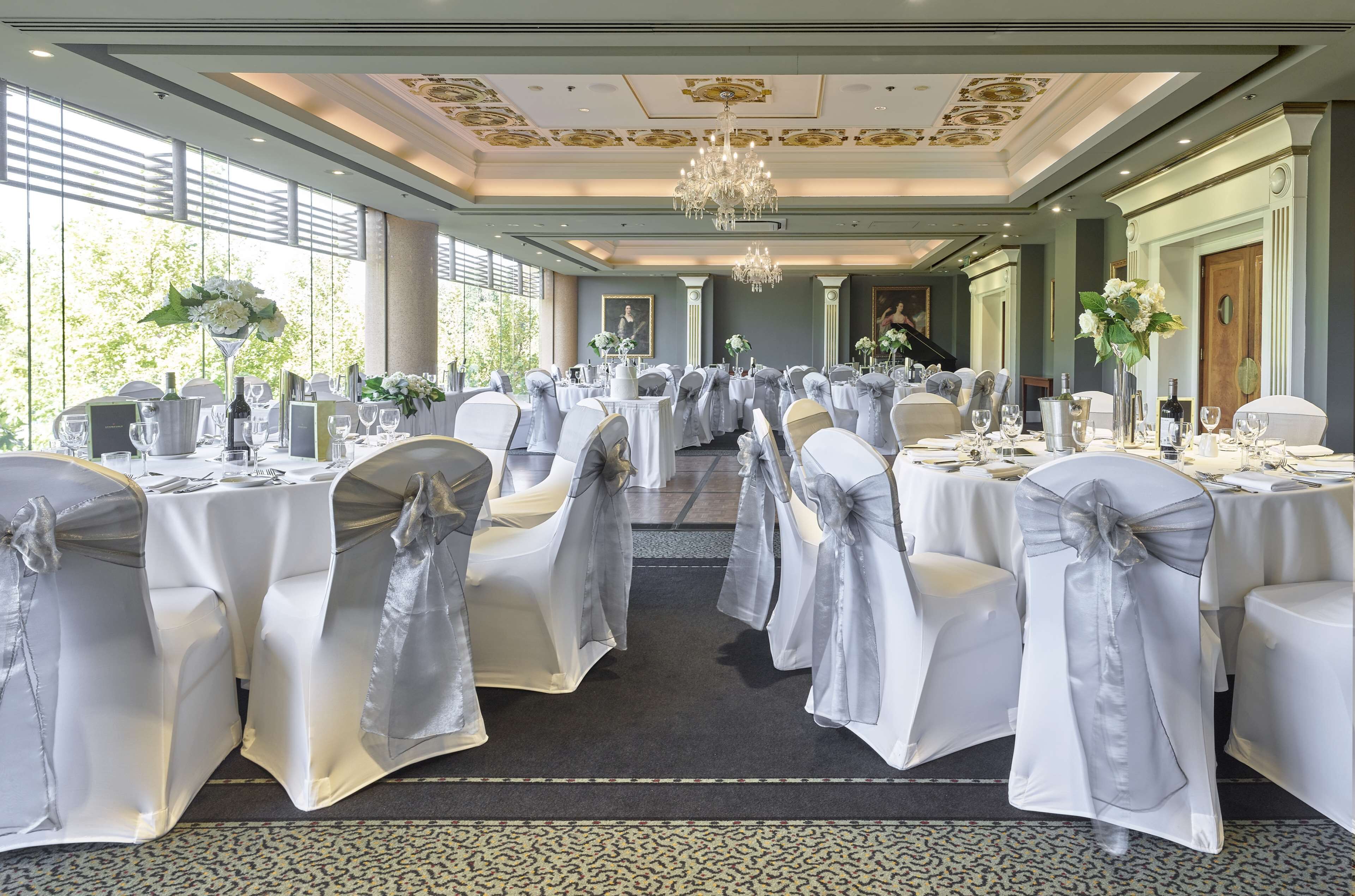 A banquet hall with round tables, chairs with white covers, chandeliers, and large windows.