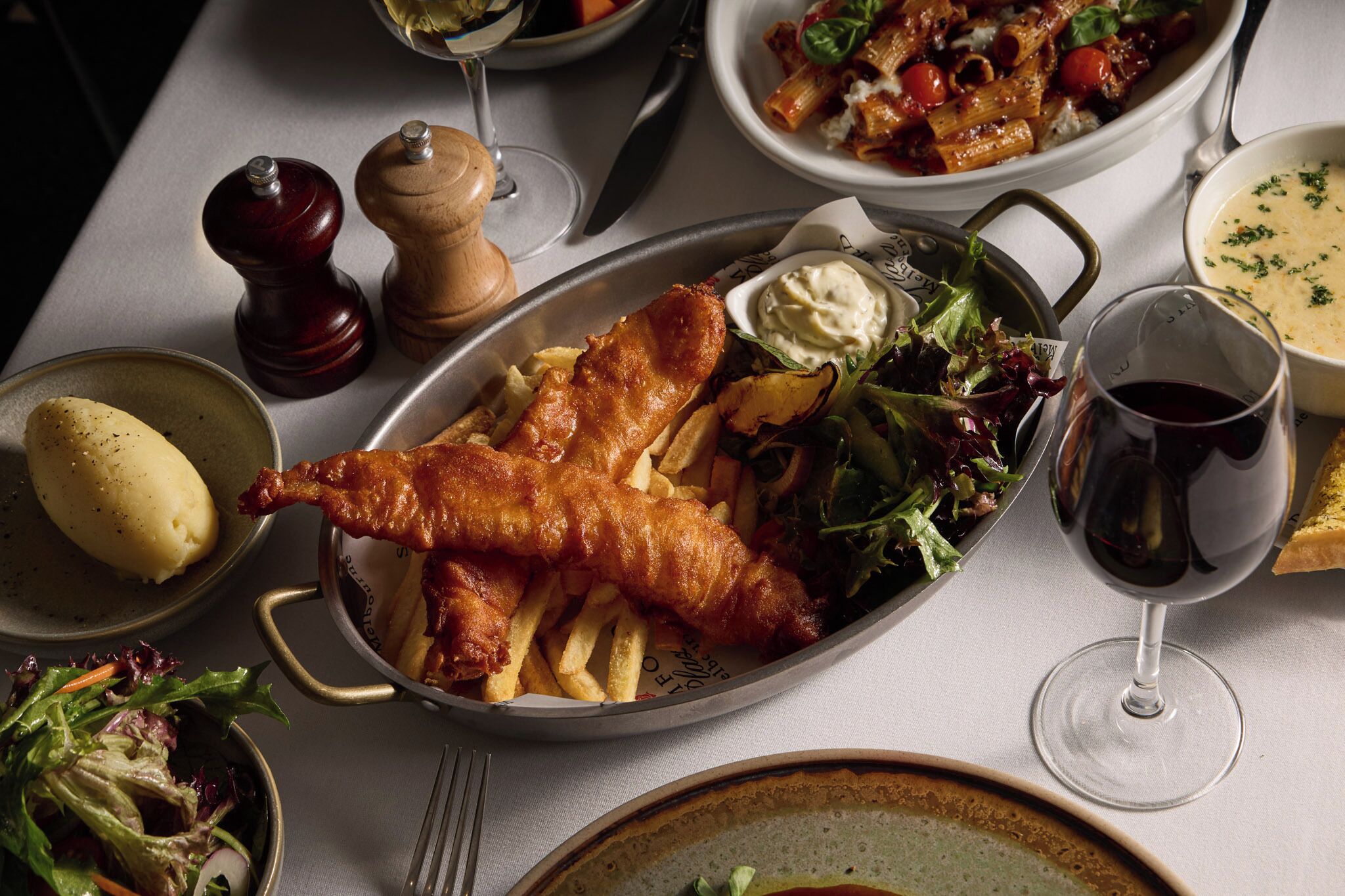 Un tavolo da pranzo con diversi piatti, tra cui pesce, patatine fritte, insalata e un bicchiere di vino rosso.