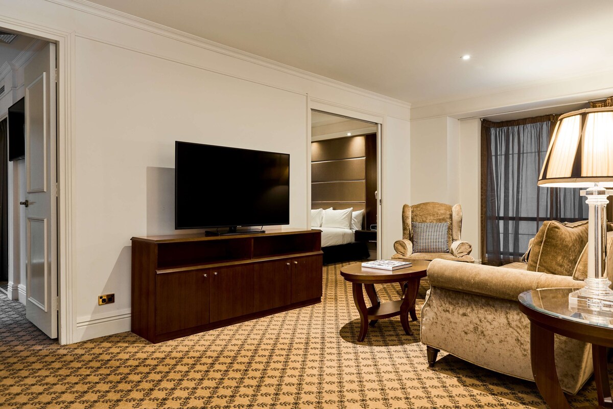 A hotel room with a television, a sofa, a chair, a bedside table, and a lamp.