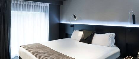 Et moderne hotelværelse med en stor seng, et natbord, en lampe og et vindue med gardiner.