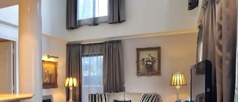 A spacious living room with a high ceiling, a sofa set, two lamps, a framed picture, and a large window with curtains.