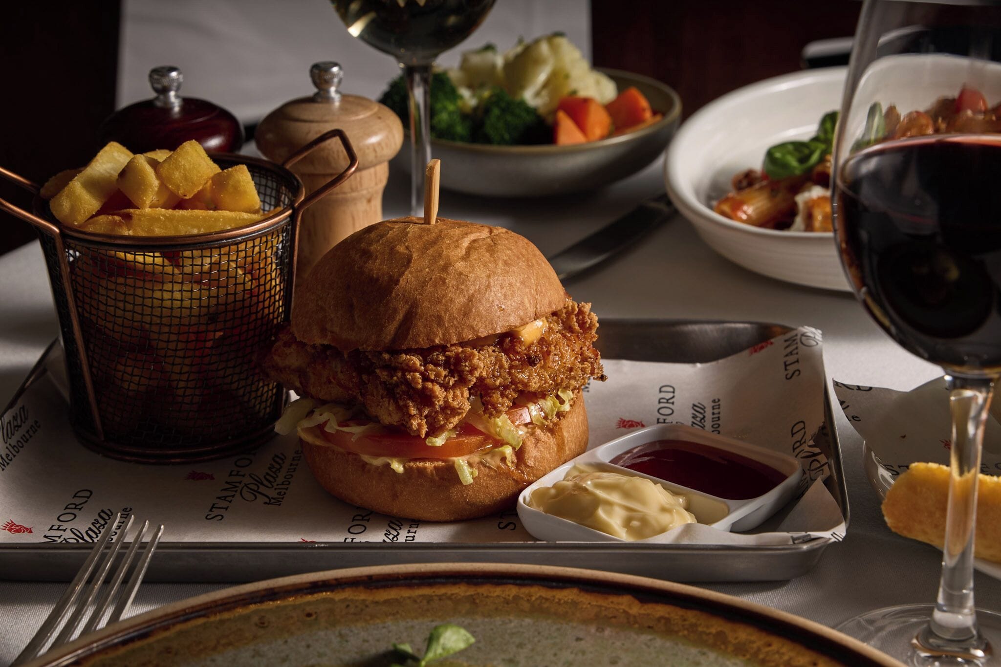 A meal with a fried chicken sandwich, fries, and a glass of red wine on a table.