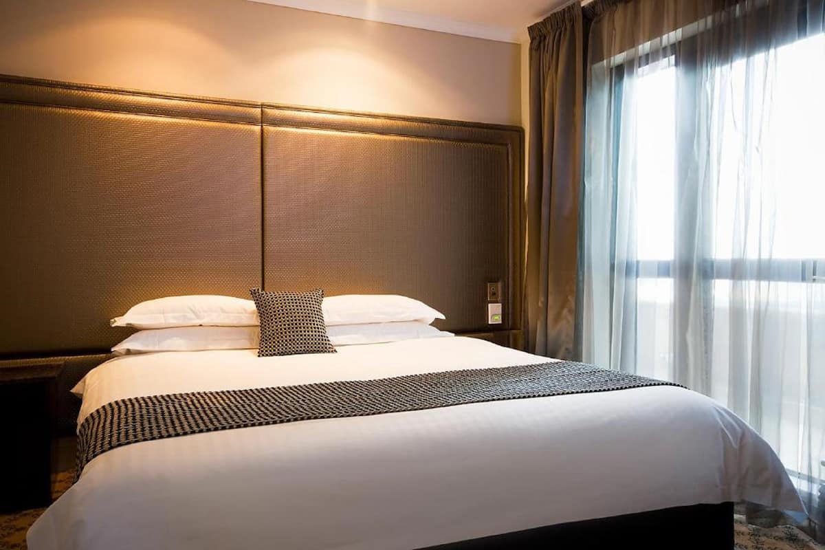 Un lit bien fait, avec du linge de lit blanc et un couvre-lit à motifs, une tête de lit au design texturé et une fenêtre ornée de rideaux légers laissant filtrer la lumière naturelle.
