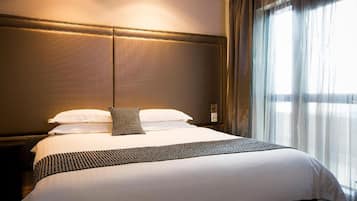 A neatly made bed with white linens and a patterned throw, a headboard with a textured design, and a window with sheer curtains allowing natural light.