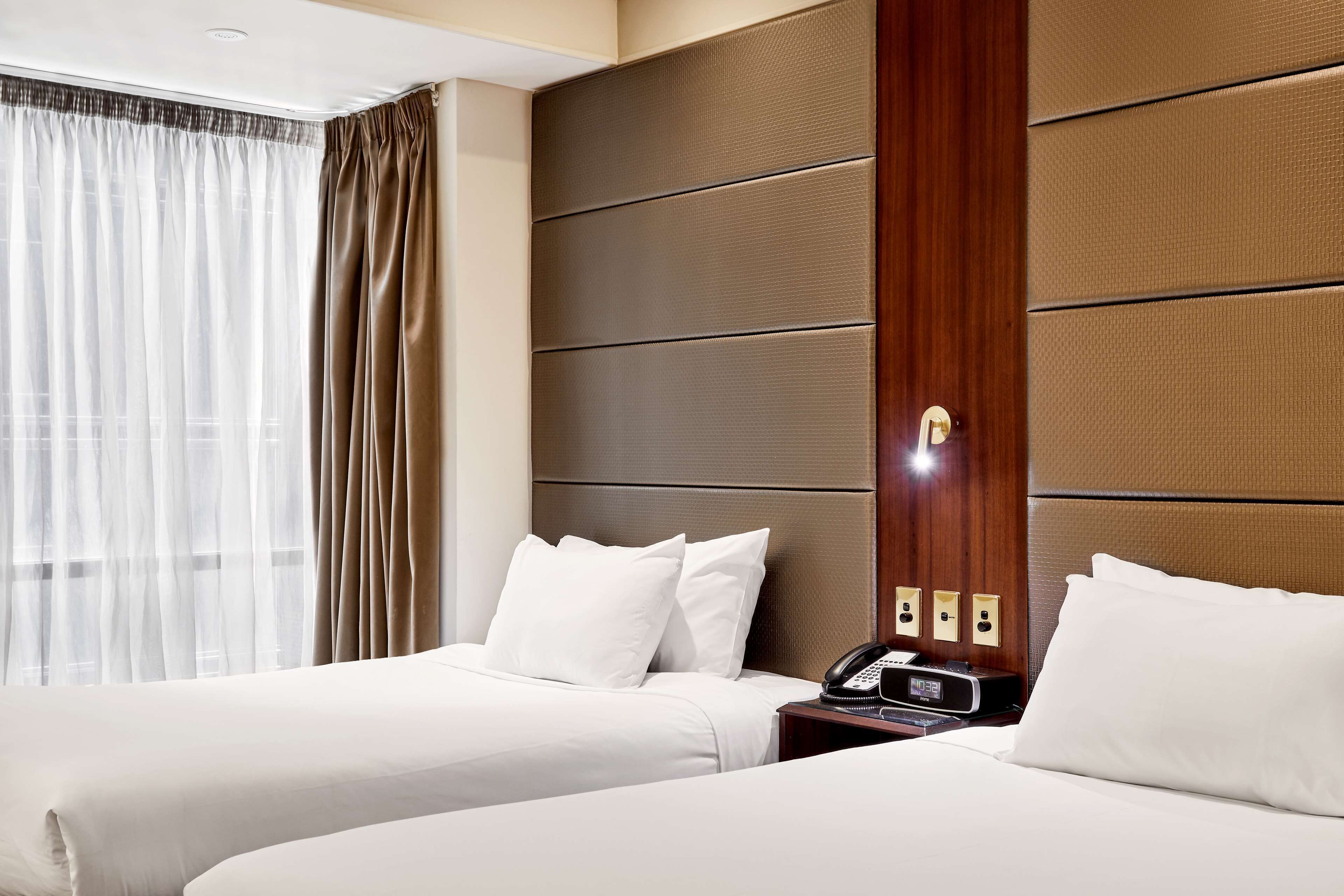 A hotel room with two beds, a wooden headboard, a bedside table with a phone, and a window with curtains.