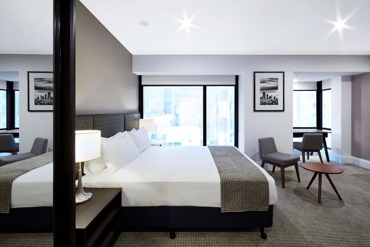 Une chambre d’hôtel avec un grand lit, deux fauteuils, une petite table et une vue sur la ville.