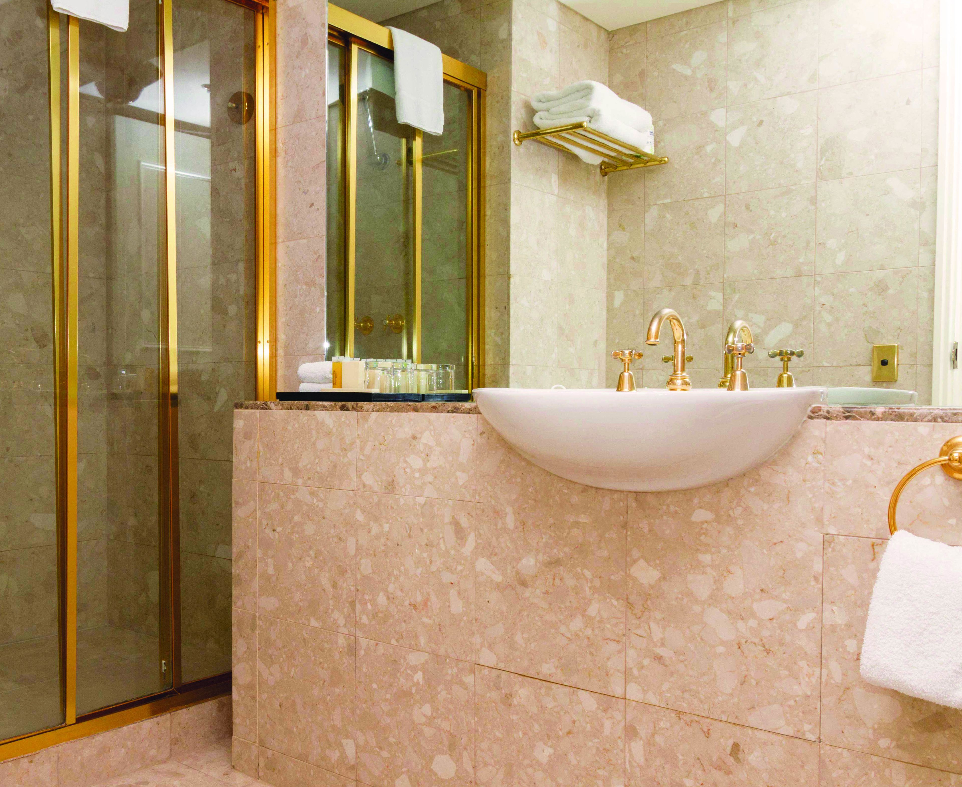 A bathroom with a sink, gold faucets, a glass shower enclosure, and marble walls.
