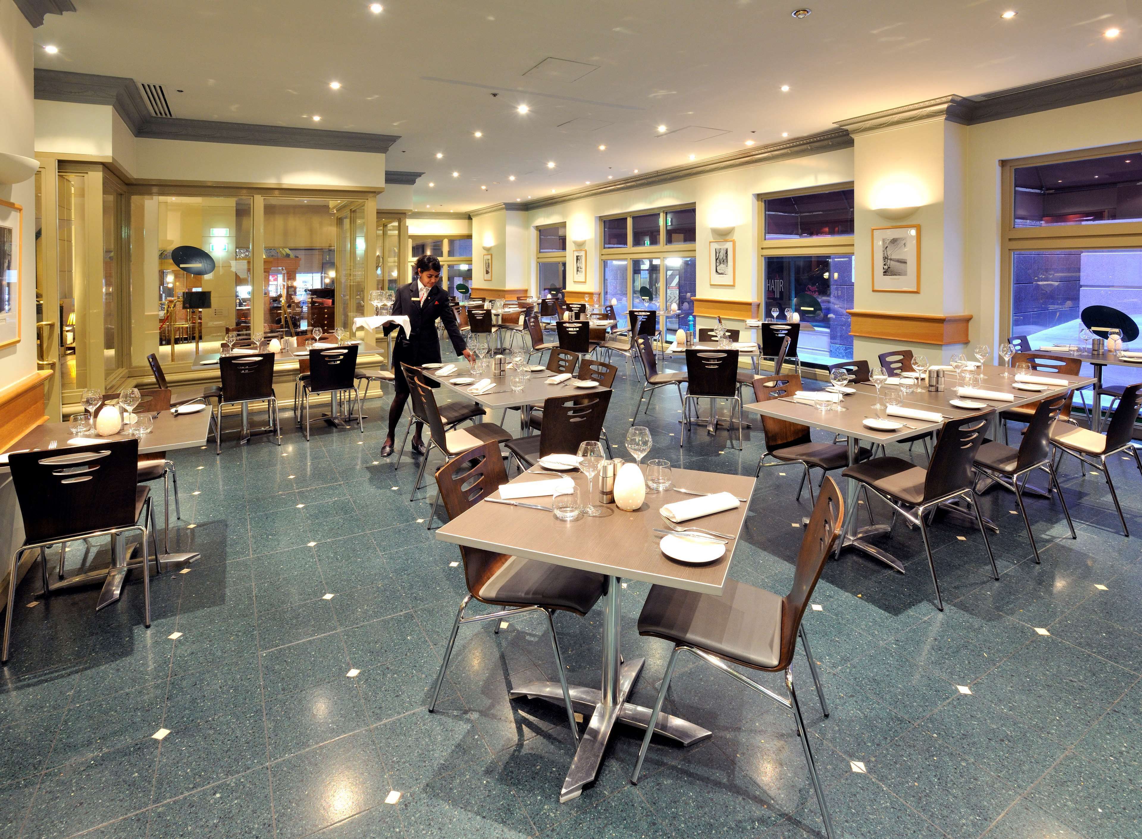 Un restaurant spacieux, doté de nombreuses tables et chaises, avec un serveur en uniforme et de grandes fenêtres laissant entrer la lumière naturelle.