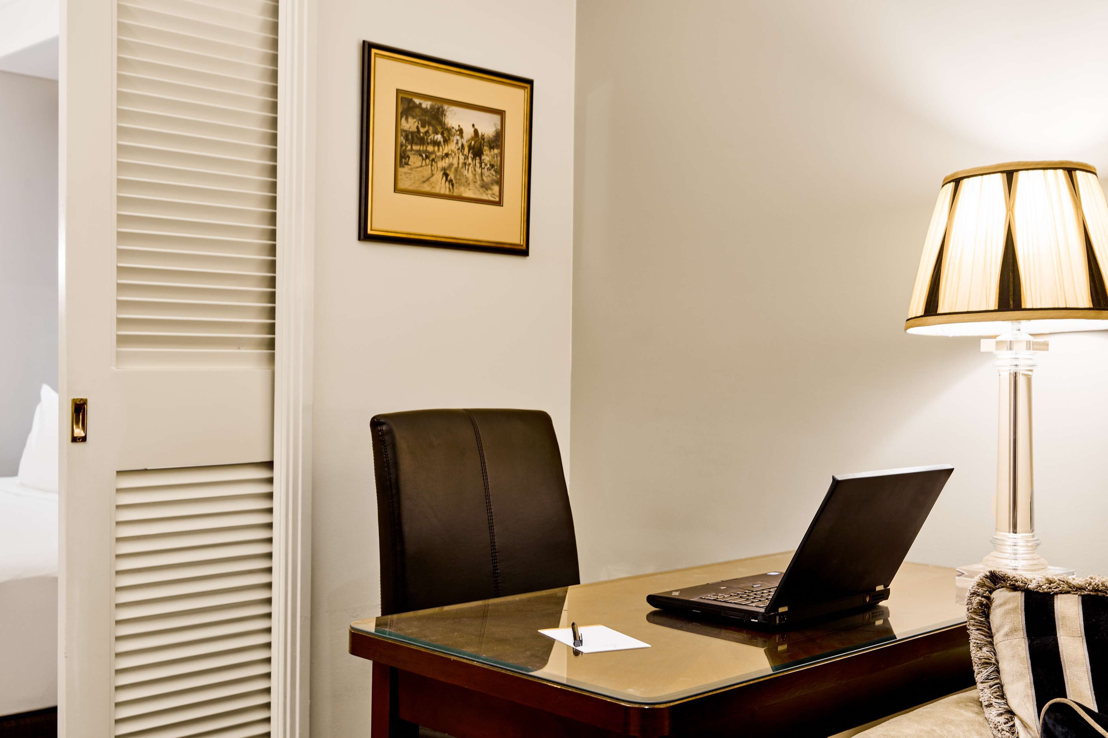 A hotel room with a desk, laptop, chair, lamp, and a framed picture on the wall.
