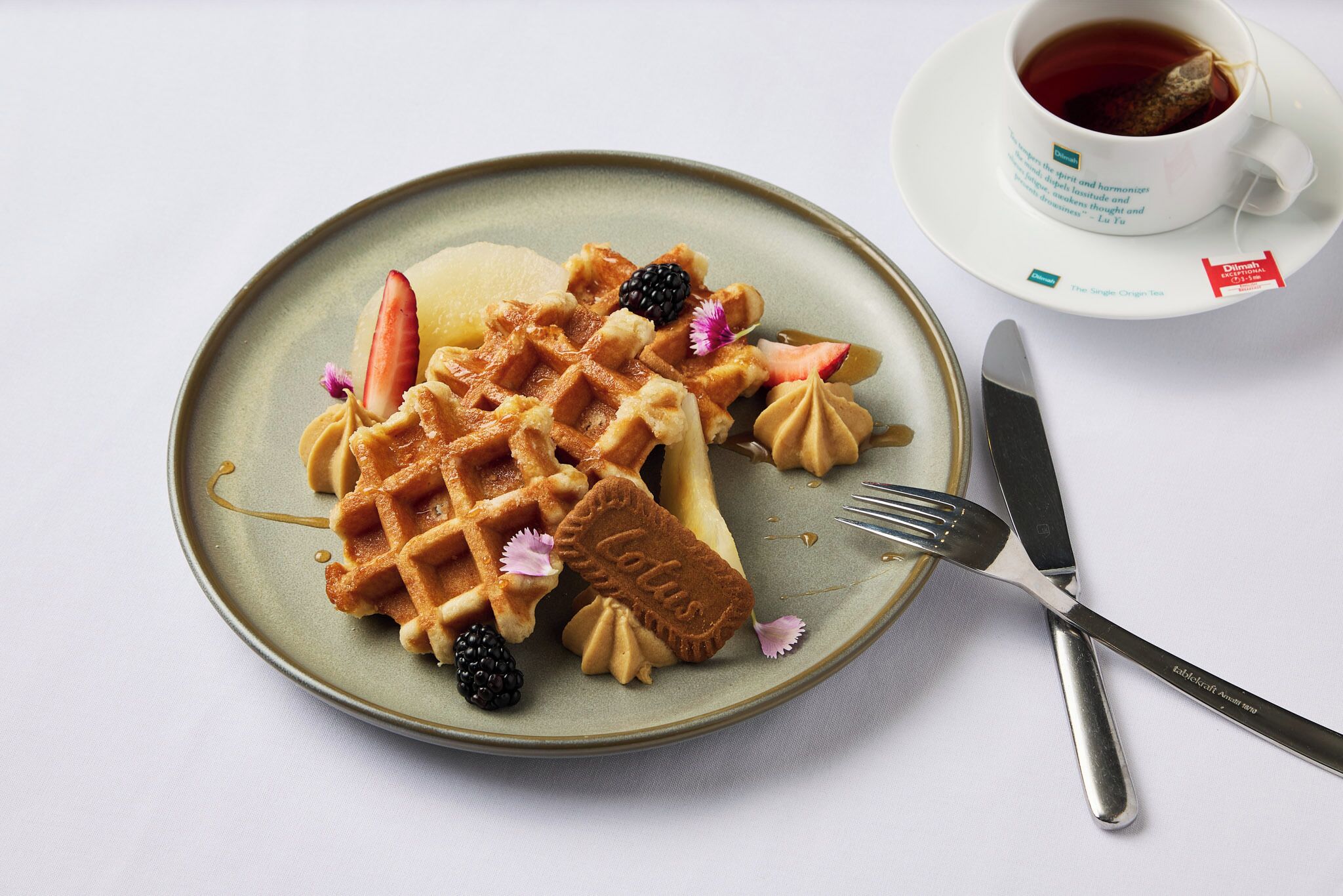 A plate with waffles, strawberries, blackberries, and a small cake, accompanied by a cup of tea and cutlery.