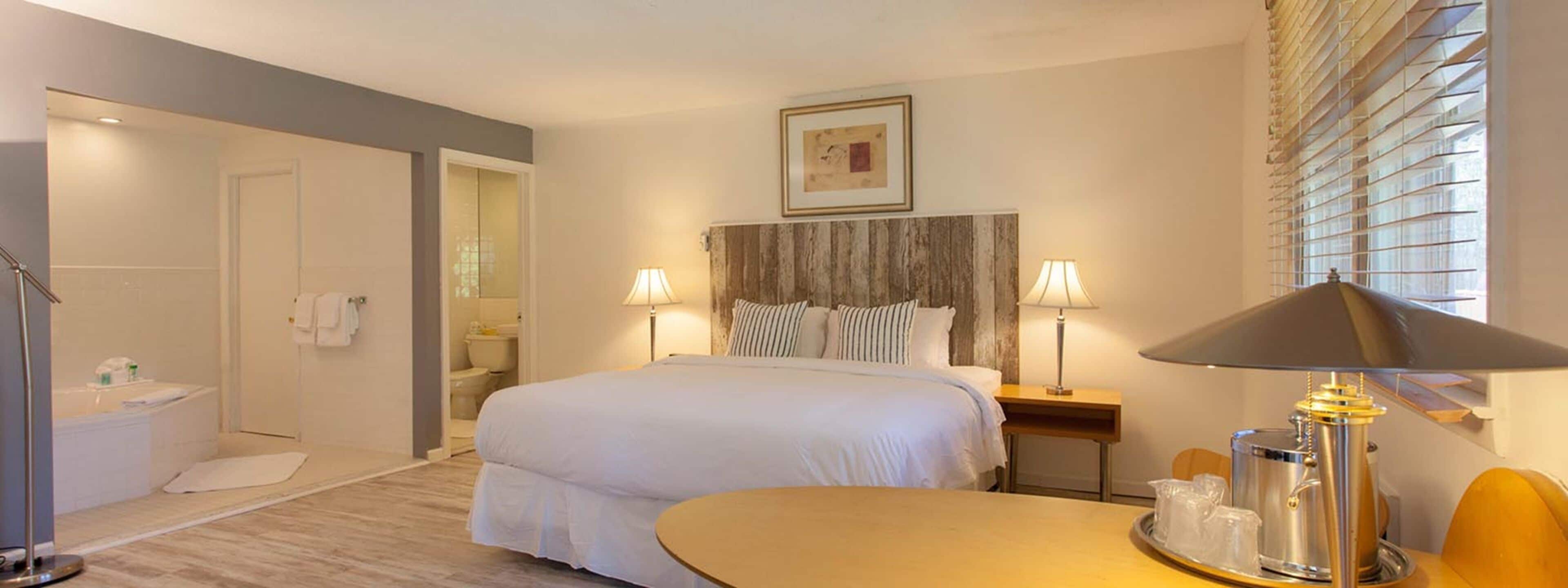 A hotel room with a bed, bedside lamps, a bedside table with a tray of glasses, a shower area, and a framed picture on the wall.