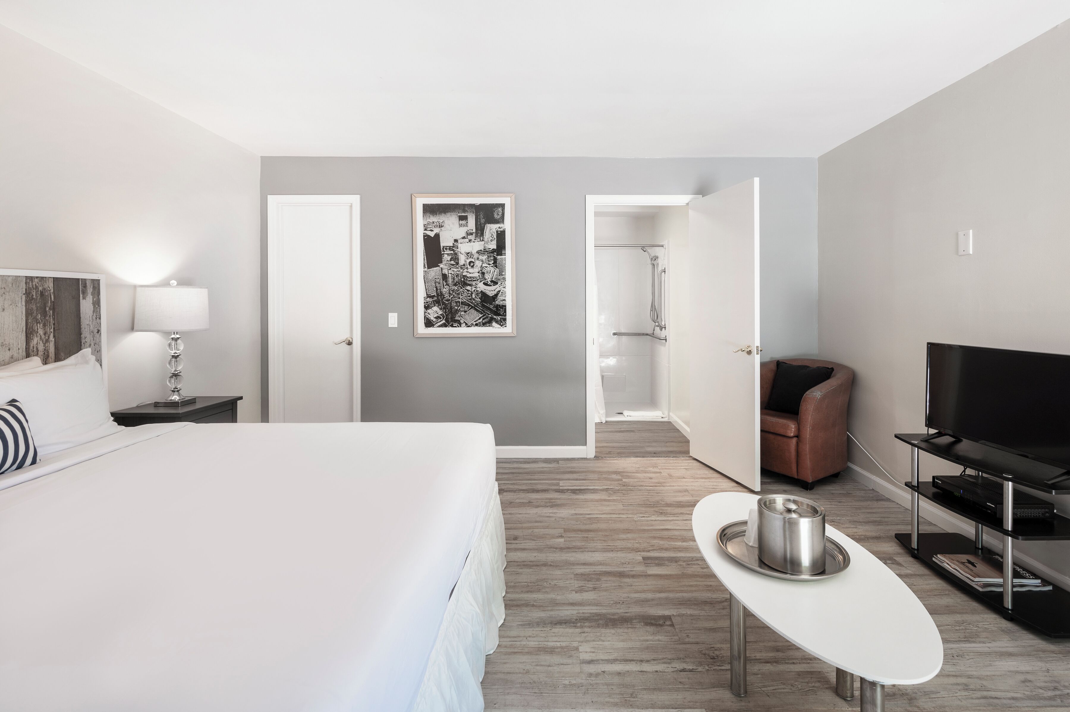A hotel room with a bed, a TV, a coffee table, a bathroom with a shower, and a door leading to another room.