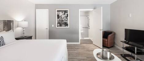 A hotel room with a bed, a TV, a coffee table, a bathroom with a shower, and a door leading to another room.