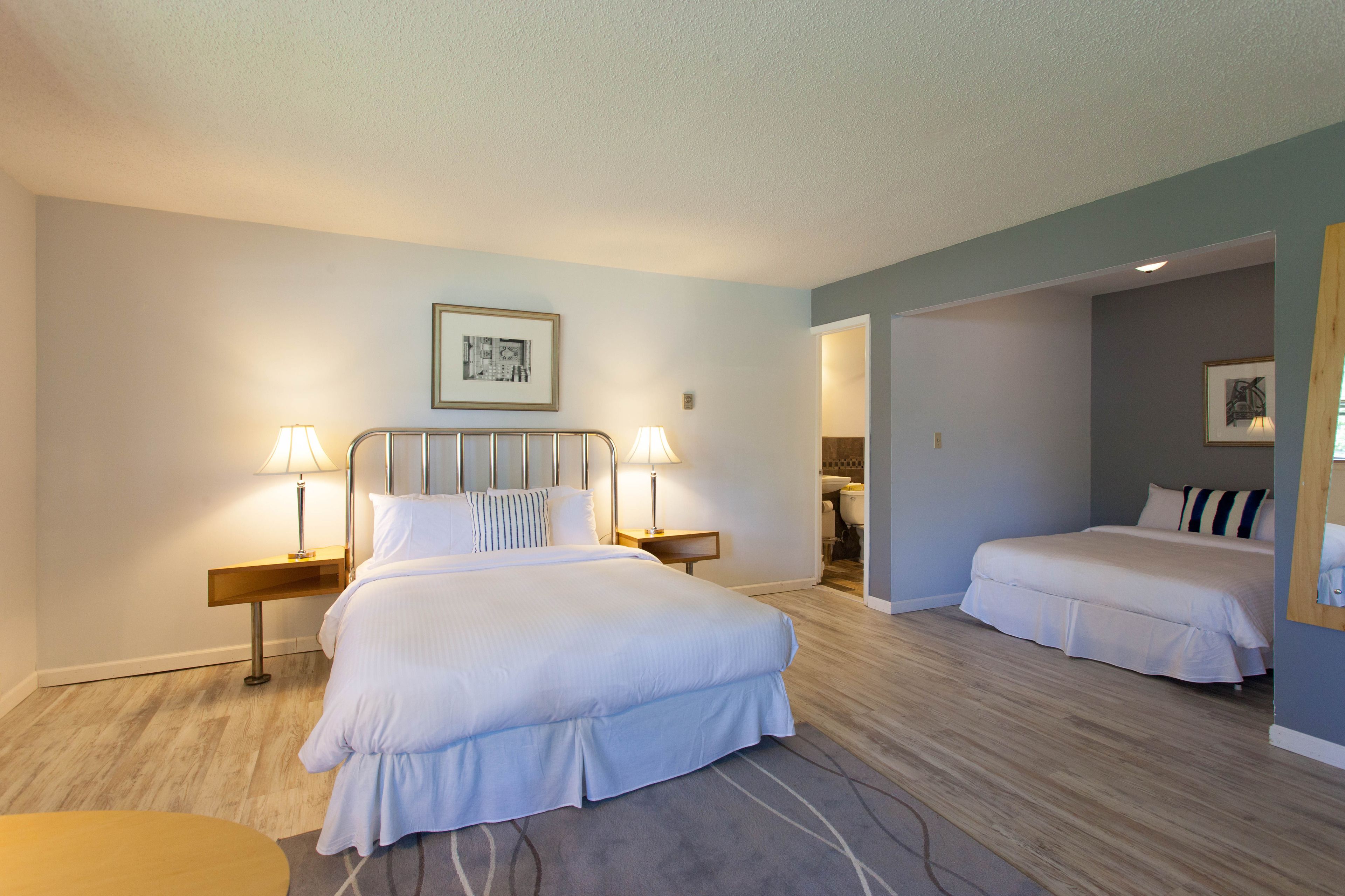 Una habitación de hotel con una cama grande, dos mesitas de noche con lámparas, un cuadro en la pared y otra cama en la habitación contigua.