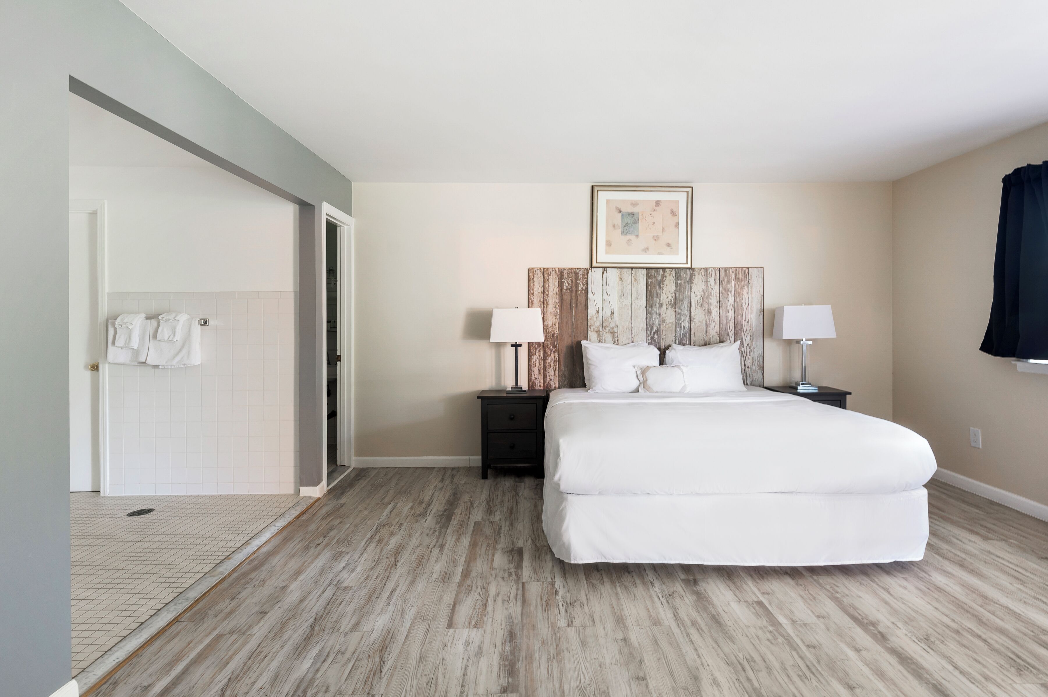 Un dormitorio con una cama grande, dos mesitas de noche con lámparas, un cabecero de madera, un cuadro enmarcado sobre la cama y un baño con tendedero.
