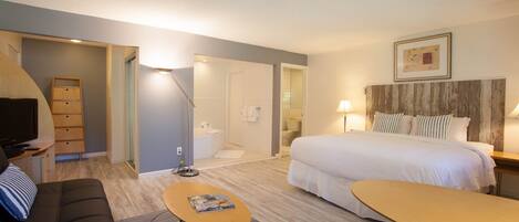 A hotel room with a bed, a sofa, a small table, a TV, and a bathroom visible through an open door.