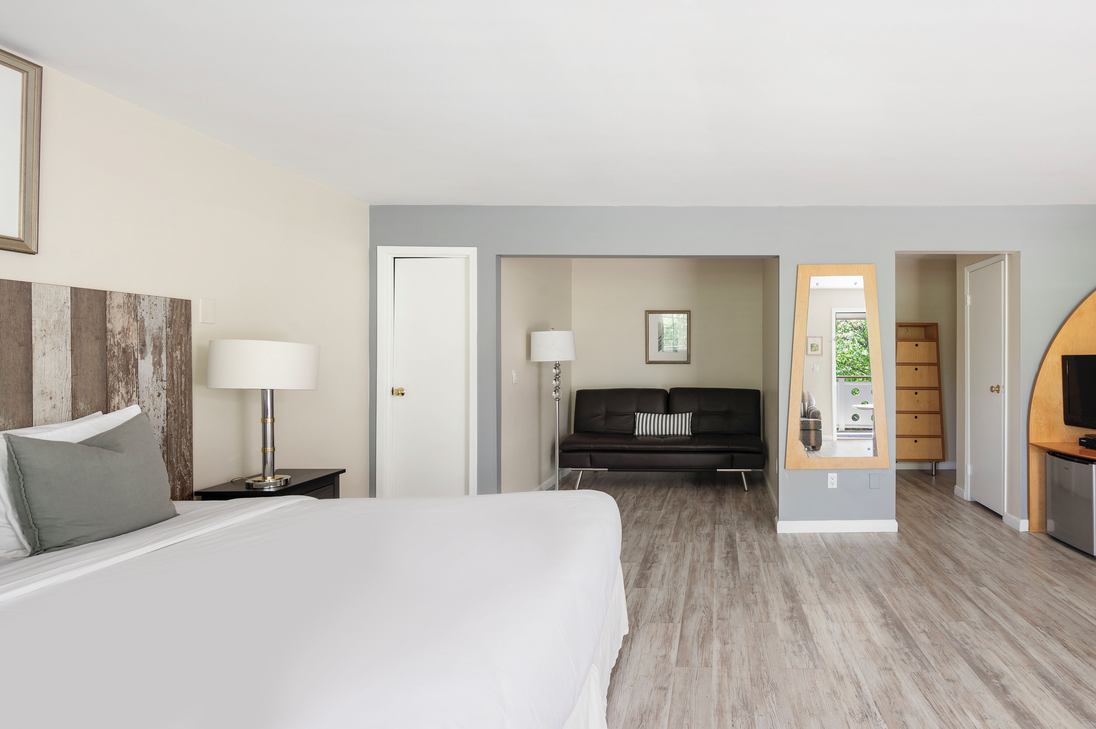 A hotel room with a bed, bedside table, lamp, and a view into the living area.