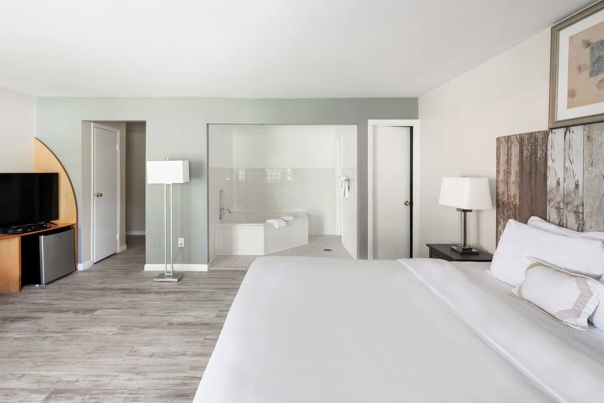A hotel room with a bed, a TV, a desk, a lamp, a bathroom with a bathtub, and a wooden accent wall.