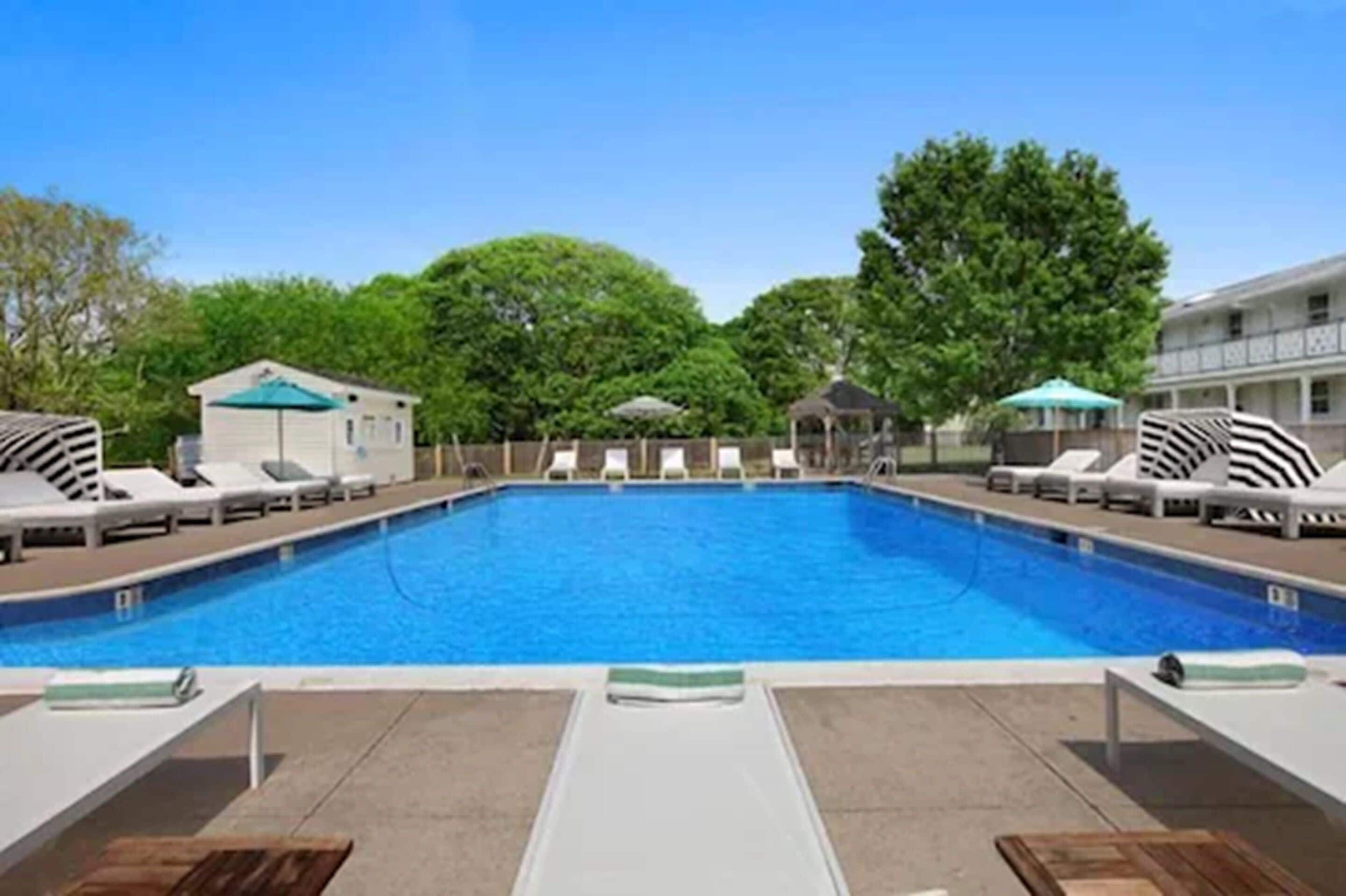 A swimming pool with lounge chairs and umbrellas, surrounded by trees and a building.