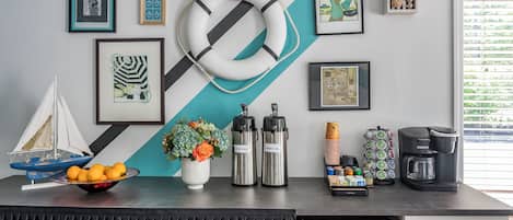 A counter with a life preserver, framed artwork, a small sailboat model, a bowl of oranges, thermos flasks, a coffee maker, and a refrigerator.