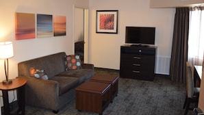 A living room with a sofa, ottomans, a TV, a lamp, and artwork on the wall. - Staybridge Suites Myrtle Beach - West by IHG (Myrtle Beach)