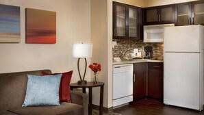 A modern living room with a sofa, a small table with a lamp, and a vase with flowers. Adjacent is a compact kitchen with a refrigerator, microwave, and coffee maker. - Staybridge Suites Myrtle Beach - West by IHG (Myrtle Beach)