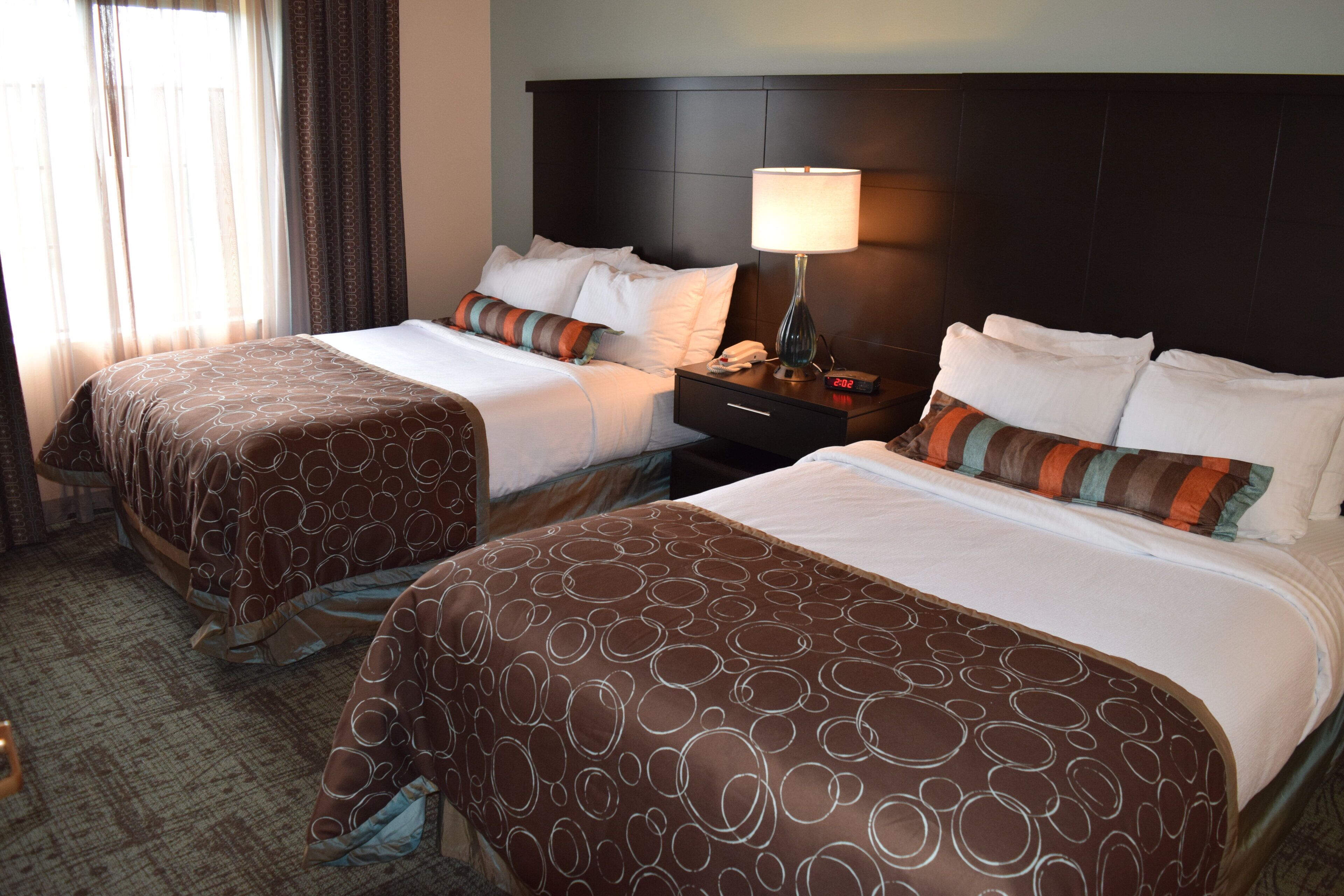 A hotel room with two beds, a nightstand with a lamp, and a window with curtains.