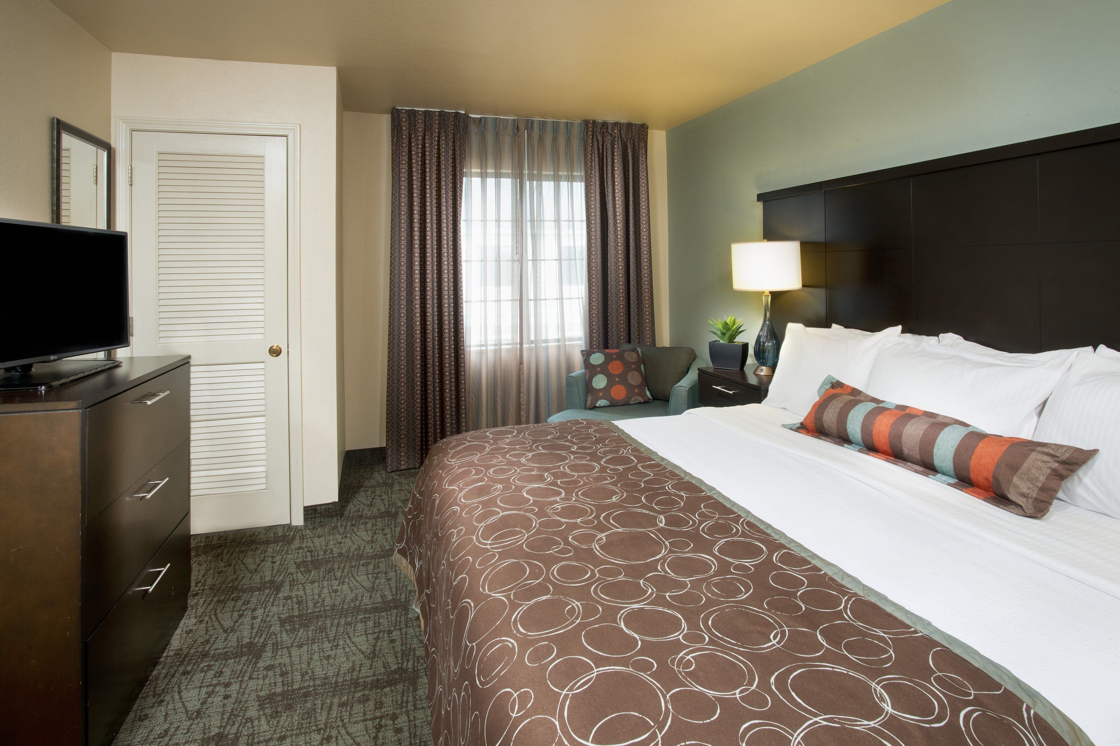 A hotel room with a large bed, a television on a dresser, a window with curtains, and a nightstand with a lamp.