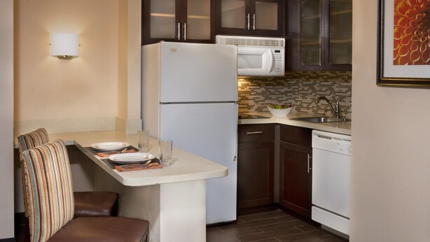 A compact kitchen with a refrigerator, microwave, and dishwasher, a small dining area with a bench, and a tiled backsplash. - Staybridge Suites Myrtle Beach - West by IHG (Myrtle Beach)