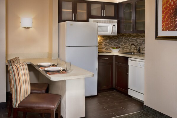 Cocina compacta con refrigerador, microondas y lavavajillas, un pequeño comedor con un banco, y salpicadero de azulejos.