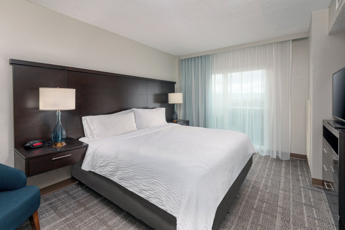 A hotel room with a large bed, two bedside tables with lamps, a TV, and a view of the outside through sheer curtains.