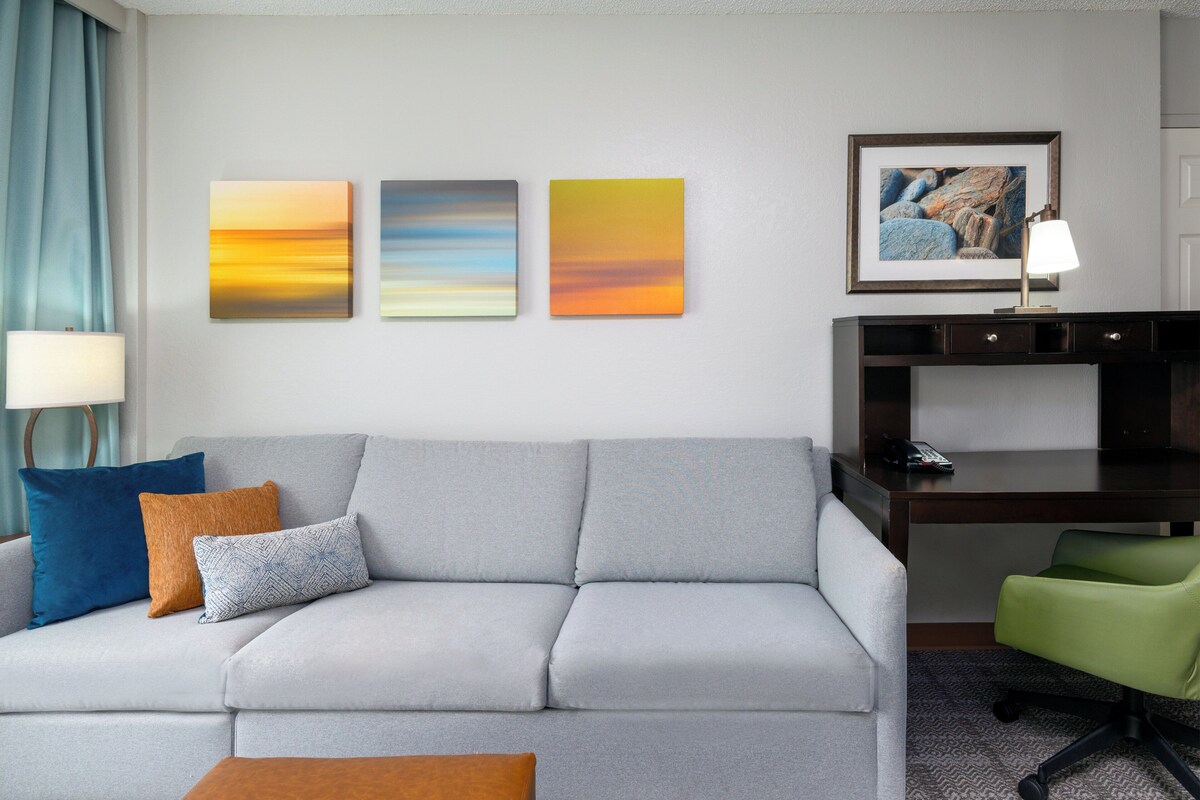 A modern hotel room with a grey sofa, a desk with a lamp, and artwork on the wall.