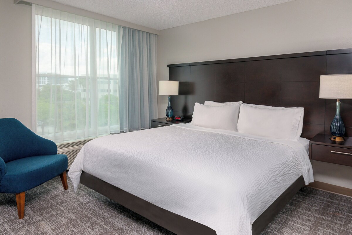 A hotel room with a large bed, two bedside lamps, a blue armchair, and a window with sheer curtains.