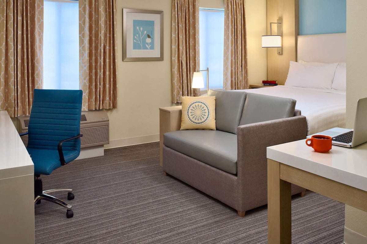 Une chambre d’hôtel avec un lit, un bureau avec un ordinateur portable, une chaise, une lampe et un tableau encadré au mur.