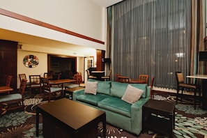 Um hall de hotel com um sofá verde, cadeiras de madeira, uma mesa de centro e uma janela grande com cortinas.