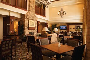 A hotel lobby with a stone fireplace, dining area with tables and chairs, a television, and a chandelier. - Staybridge Suites Irving Las Colinas by IHG (Irving)
