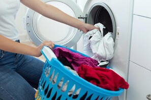 A person loading a washing machine with a blue laundry basket containing various clothes.