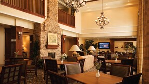 A hotel lobby with a stone fireplace, dining area with tables and chairs, a television, and a chandelier.