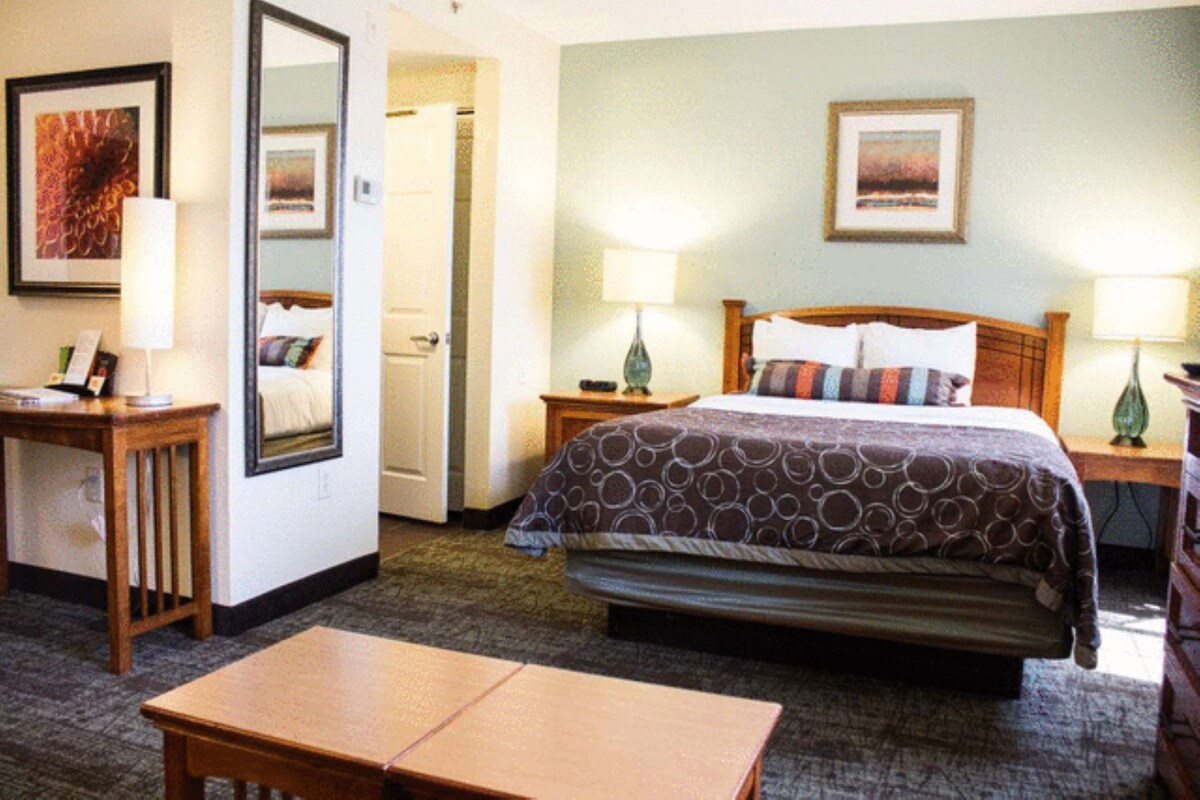 Una habitación de hotel con una cama, dos mesitas de noche, un escritorio, una silla, un espejo y una puerta.