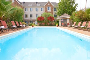 A swimming pool with lounge chairs and umbrellas, surrounded by a building with multiple windows and a stone facade. - Staybridge Suites Irving Las Colinas by IHG (Irving)
