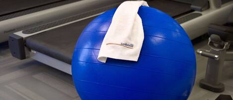 Una pelota de ejercicio azul con una toalla blanca encima, sobre el suelo de un gimnasio con una cinta de correr y mancuernas cerca.