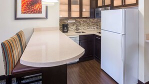 A compact kitchen with a built-in island, dark wooden cabinets, a white refrigerator, and a striped cushion on a bench. - Sonesta ES Suites Atlanta - Perimeter Center (Atlanta)