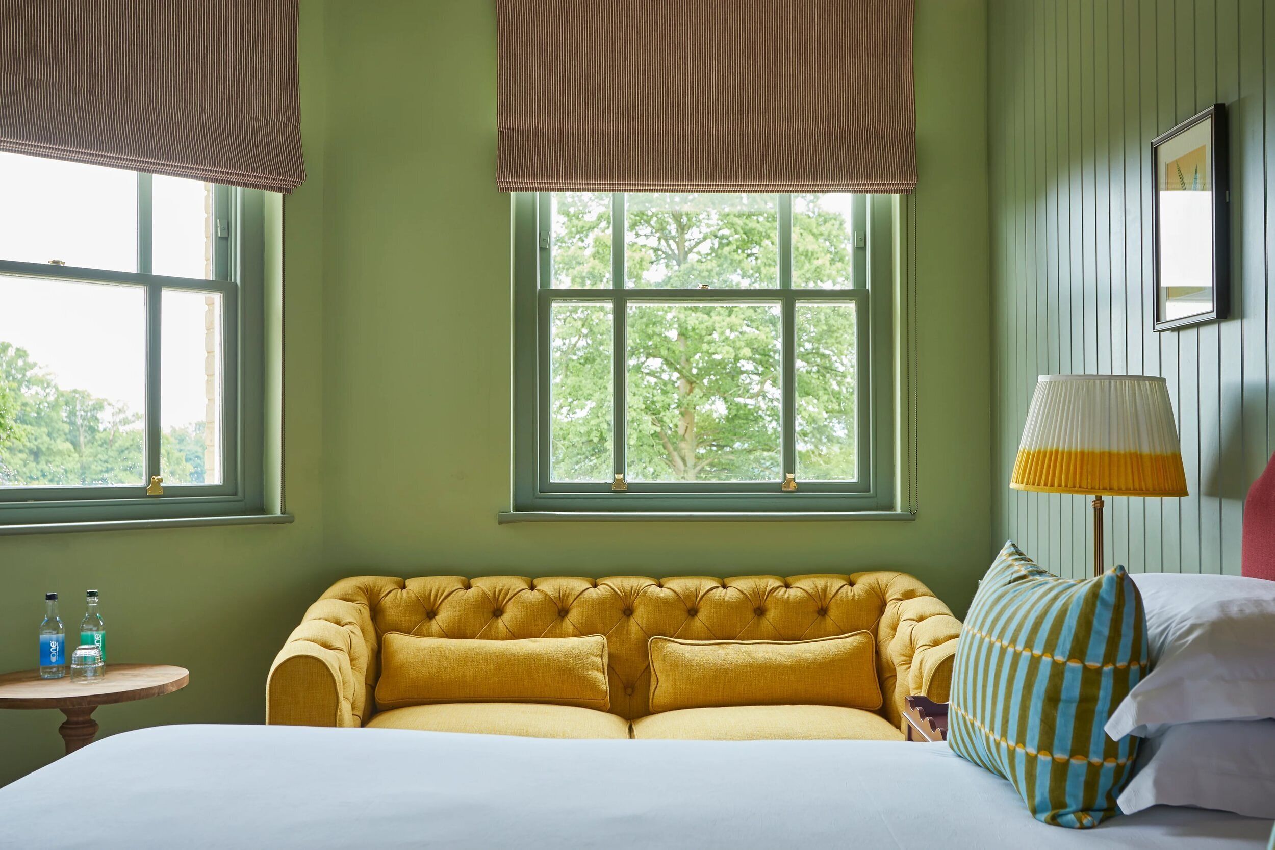 A room with a large window, a tufted yellow sofa, a bedside table with bottles, and a lamp.