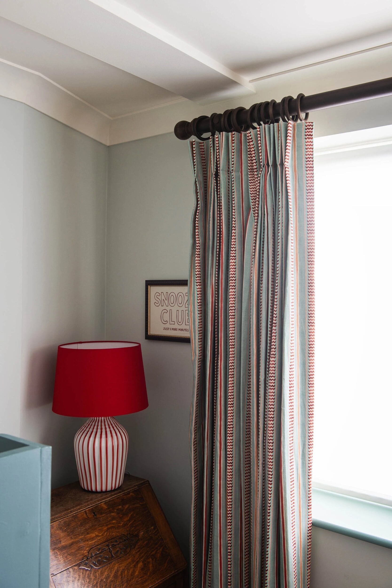 A room with a striped curtain, a red lamp on a wooden nightstand, and a framed sign on the wall.