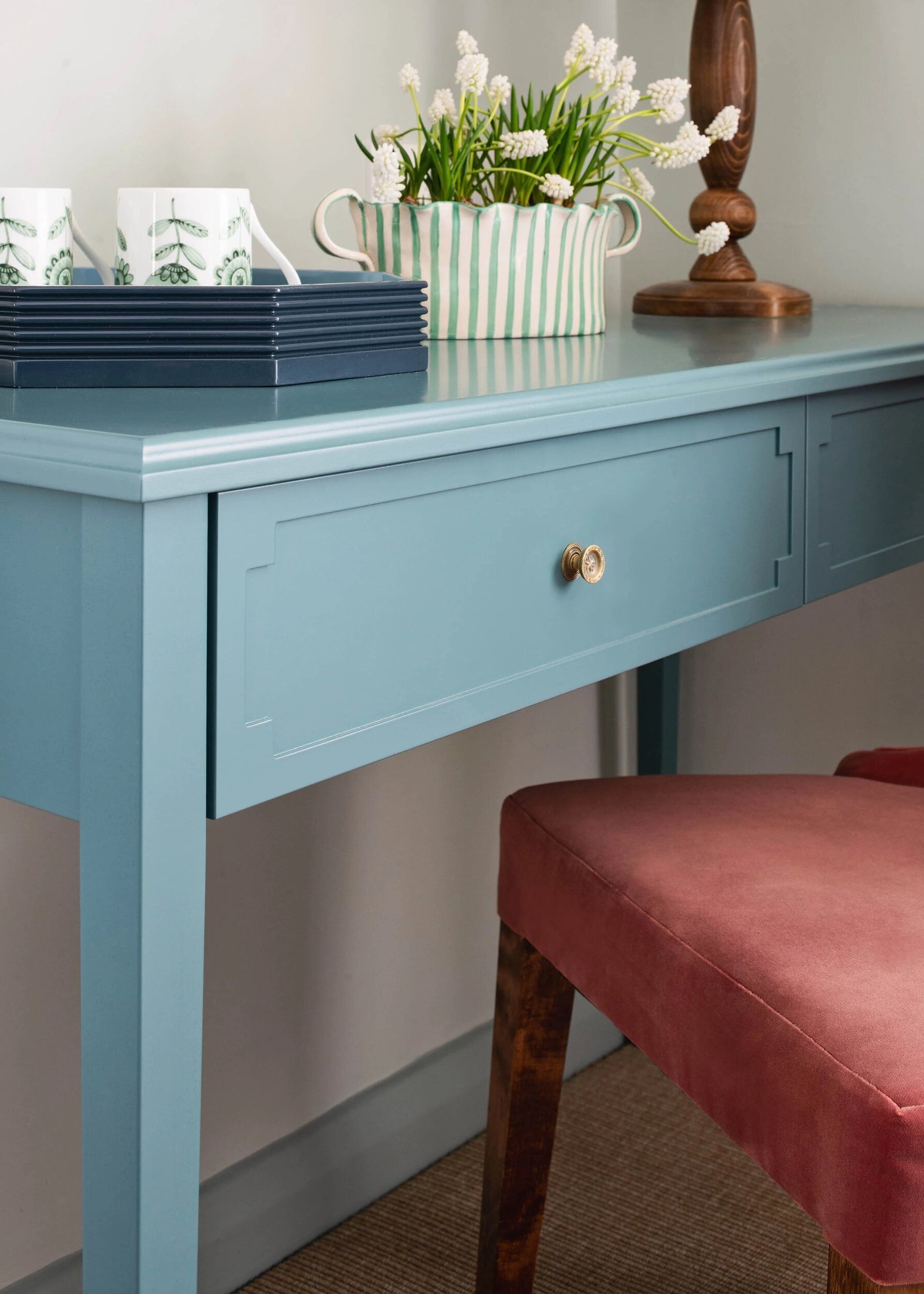 A light blue desk with a drawer, a striped vase with flowers, and a stack of blue mugs.