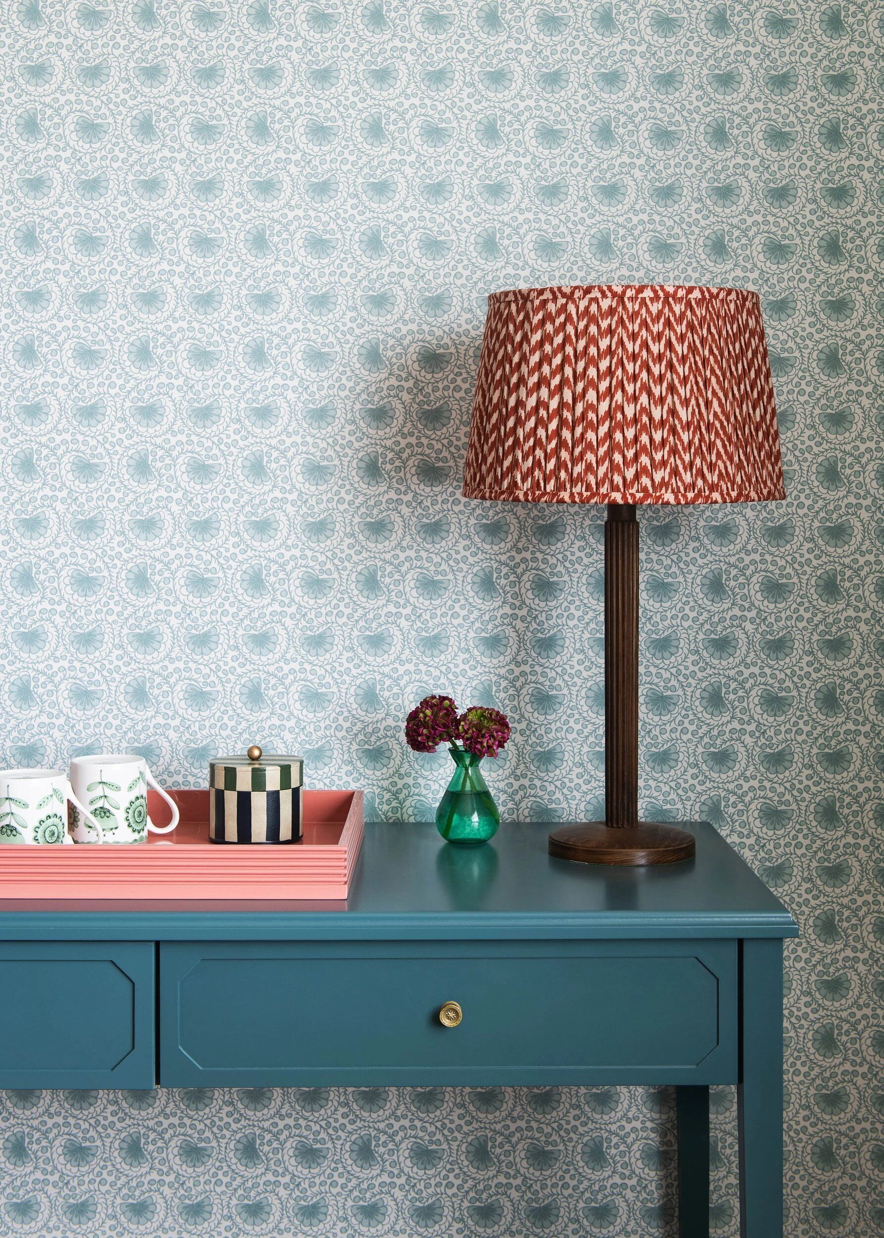 A teal-colored dresser with a drawer, a striped lamp with a patterned shade, a green vase with flowers, and two mugs with a leaf pattern.