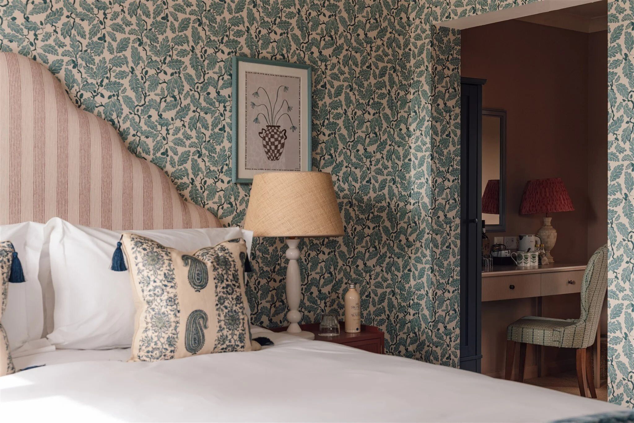 A bedroom with a bed, a nightstand, a lamp, a desk, a chair, and a mirror.