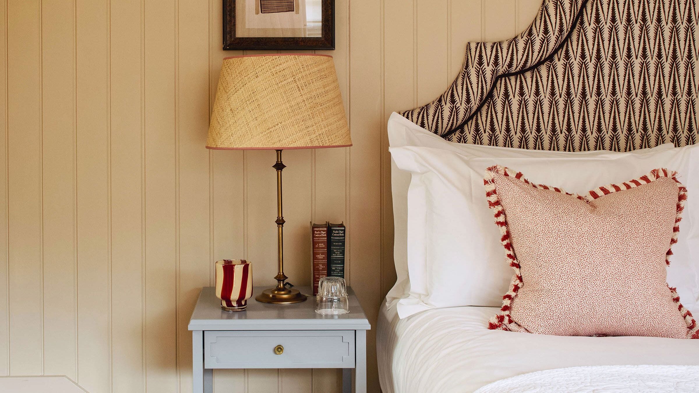 Una cama bien hecha con sábanas blancas y almohadas decorativas, una lámpara de noche, una mesita de noche con un libro y un vaso, y un cuadro en la pared.
